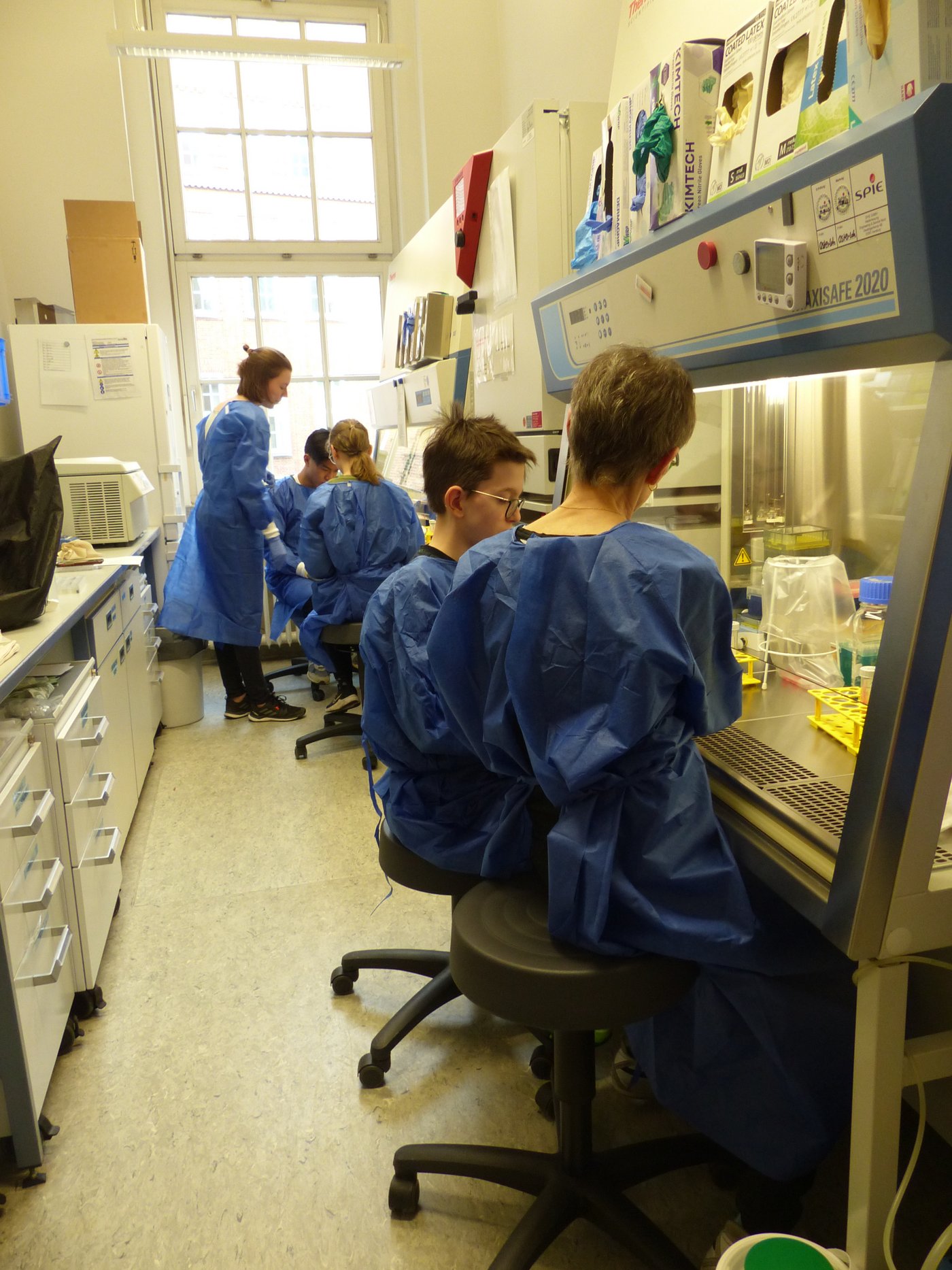Das Foto zeigt mehrere Kinder in blauen Schutzanzügen entlang zweiter Lab Benches bei der Laborarbeit.
